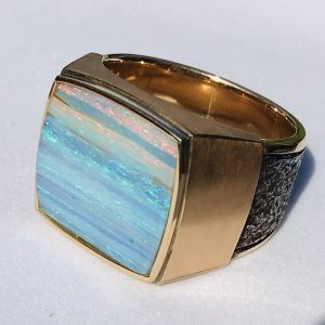 This cushion cut Blue Australian crystal opal Is full of blue and red flashes. The stone is precision cut into the bezel, (16 x 16 mm). This large men’s ring is heavy and Fabricated in 18 karat royal yellow Gold, and forged stainless steel. (size 10 3/4)